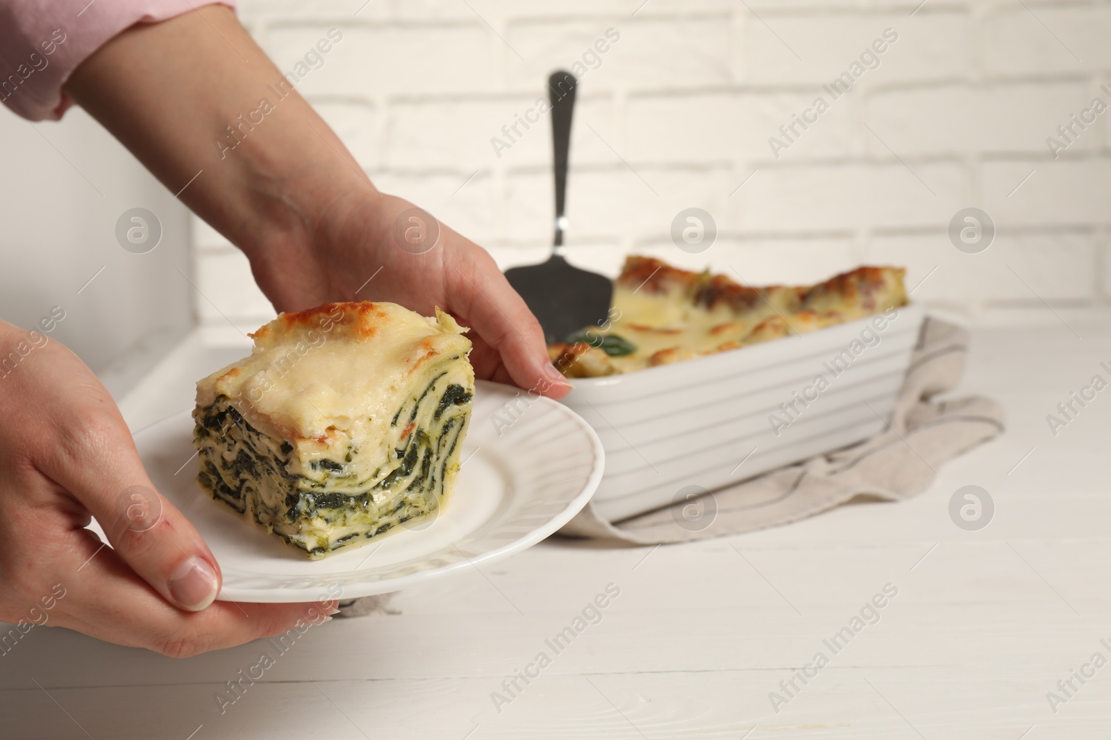 Photo of Woman holding plate with piece of delicious spinach lasagne at white wooden table, closeup