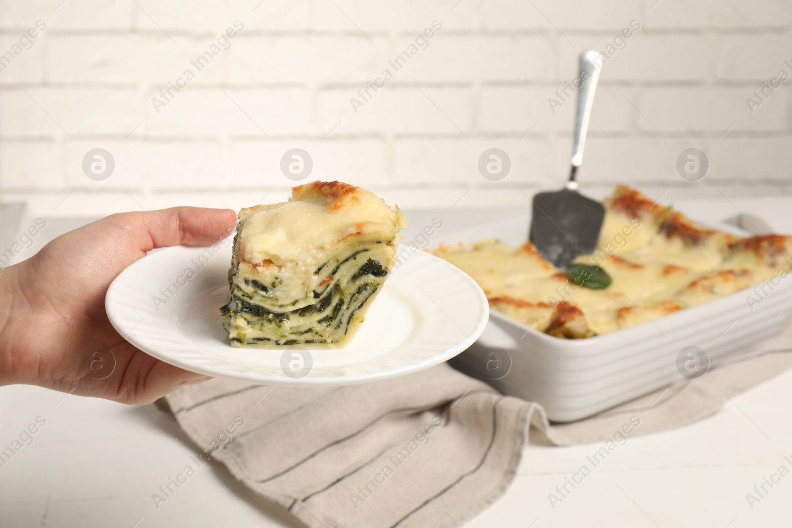 Photo of Woman holding plate with piece of delicious spinach lasagne at white table, closeup