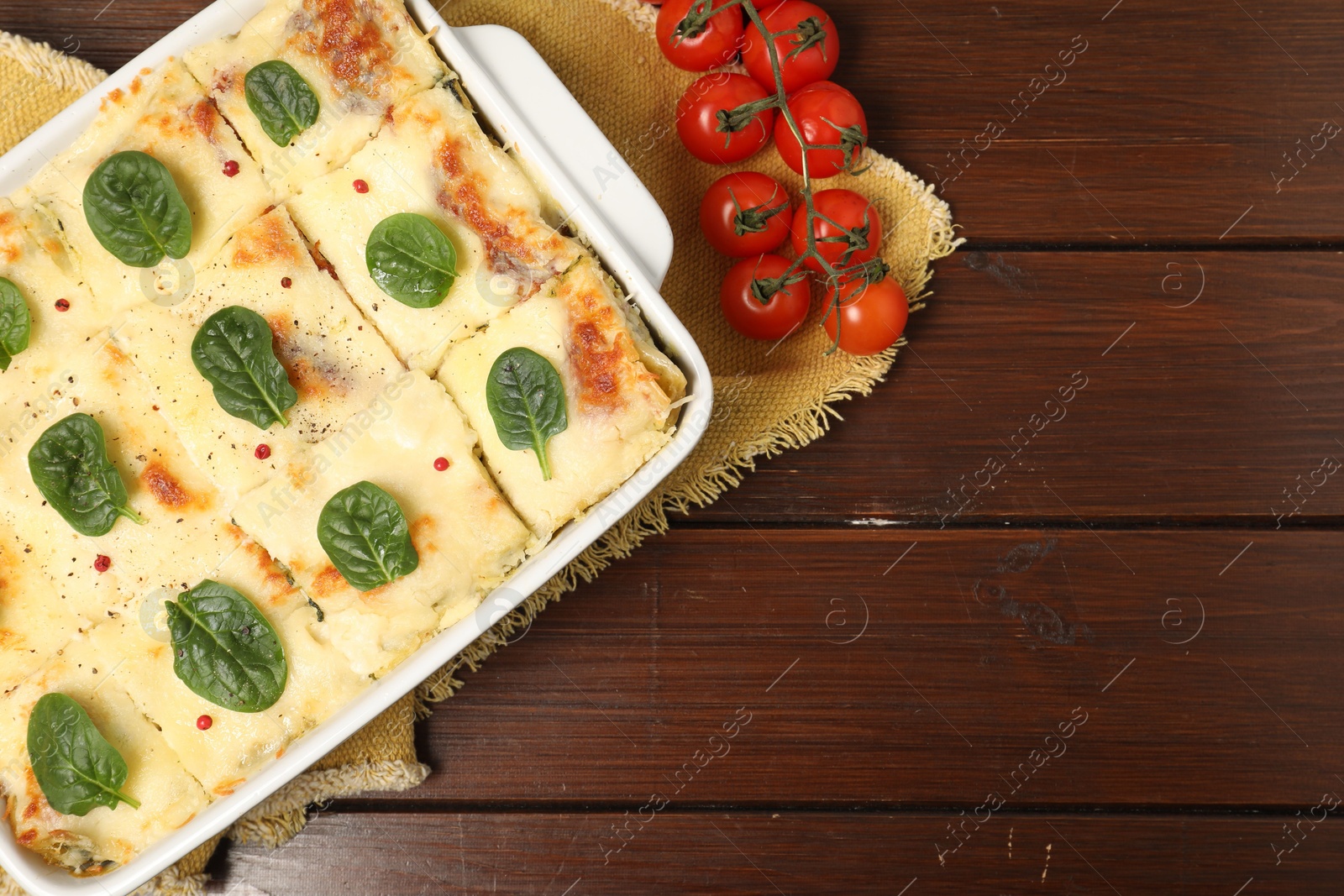 Photo of Delicious spinach lasagne in baking dish and fresh tomatoes on wooden table, flat lay. Space for text