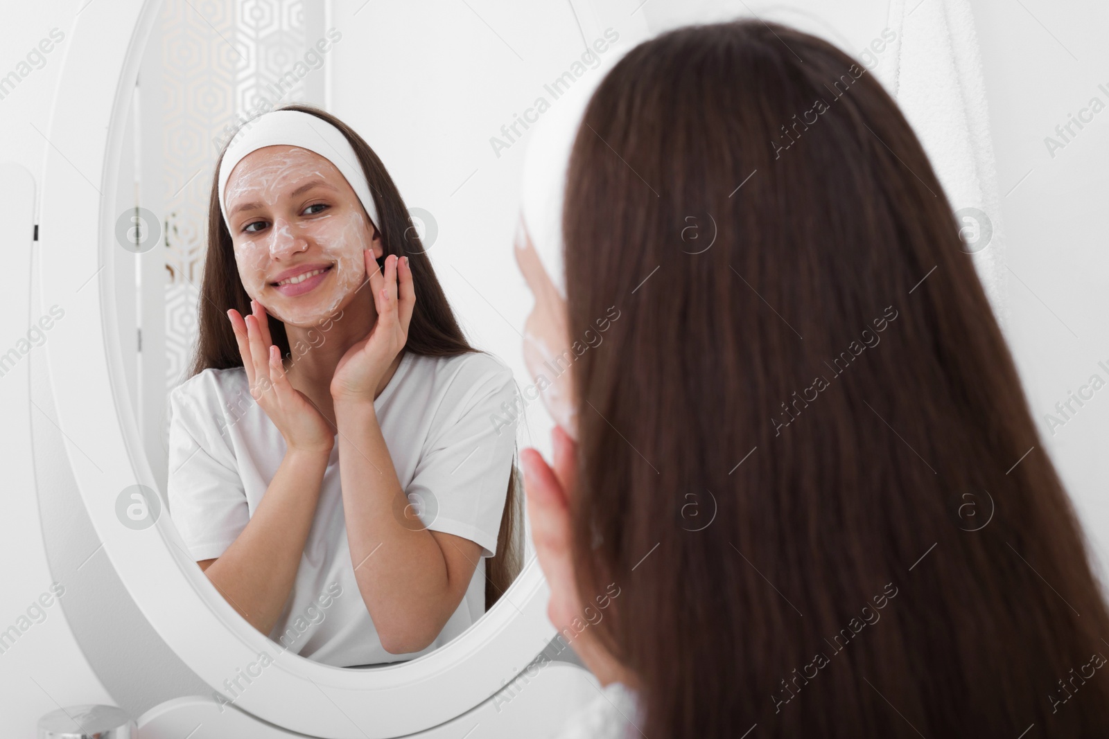 Photo of Teenage girl applying cream onto face near mirror indoors. Acne treatment