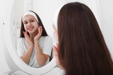 Photo of Teenage girl applying cream onto face near mirror indoors. Acne treatment