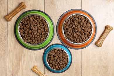 Photo of Dry pet food in feeding bowls and chew bones on wooden floor, flat lay