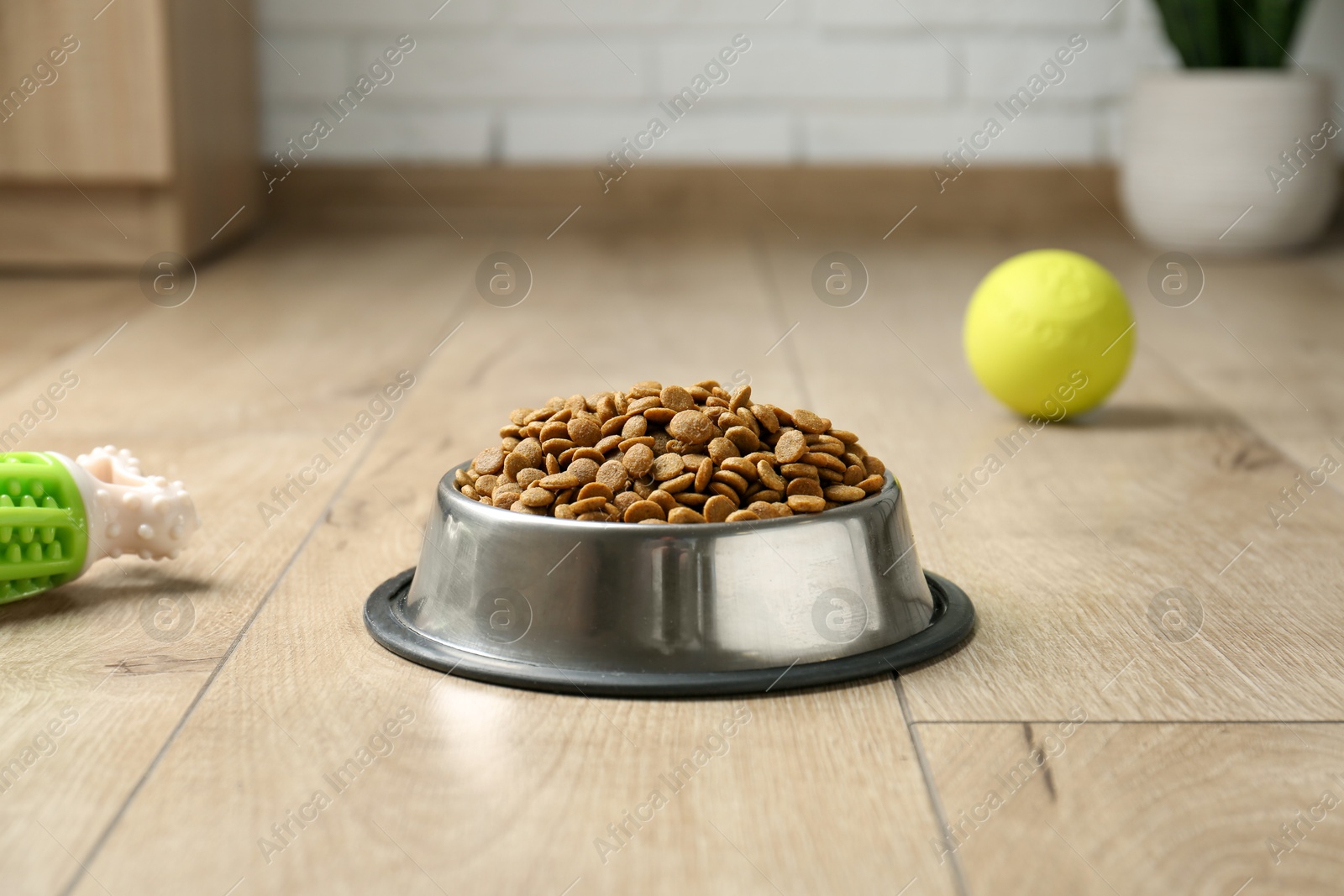 Photo of Dry pet food in feeding bowl and toys on floor indoors