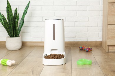 Photo of Dry pet food in automatic feeder and toys on floor indoors