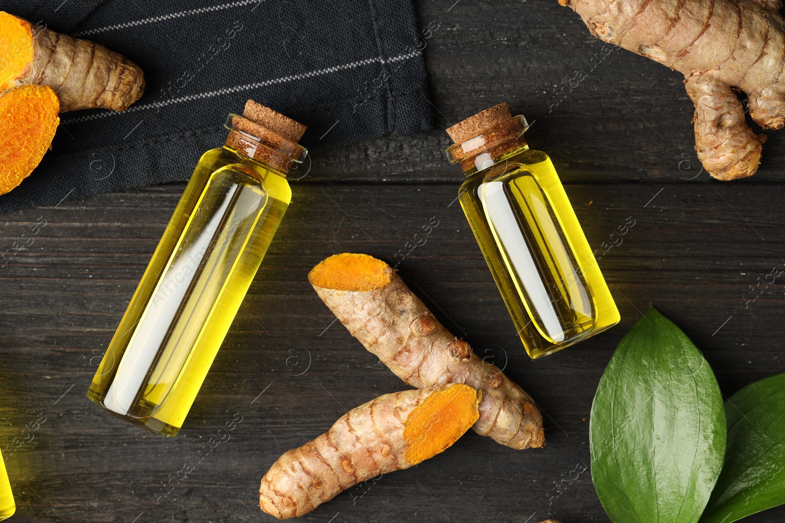 Photo of Turmeric oil, roots and green leaves on dark wooden table, flat lay