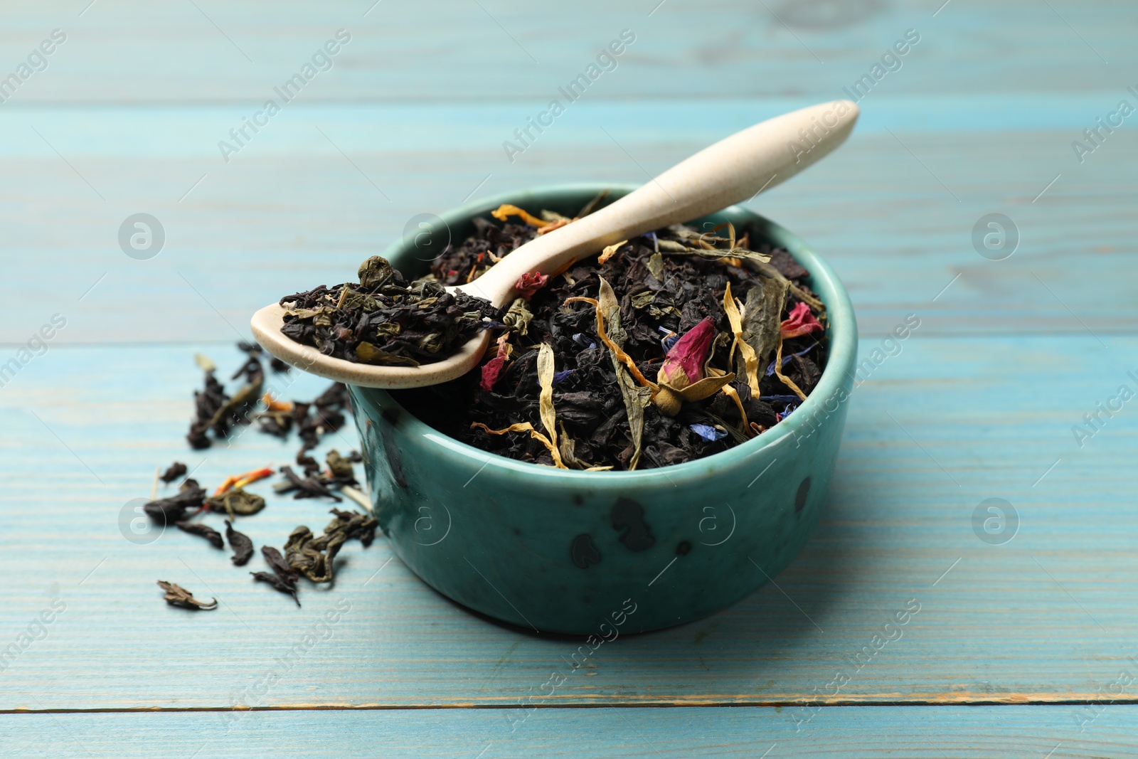 Photo of Dry tea leaves in bowl and spoon on light blue wooden table, closeup