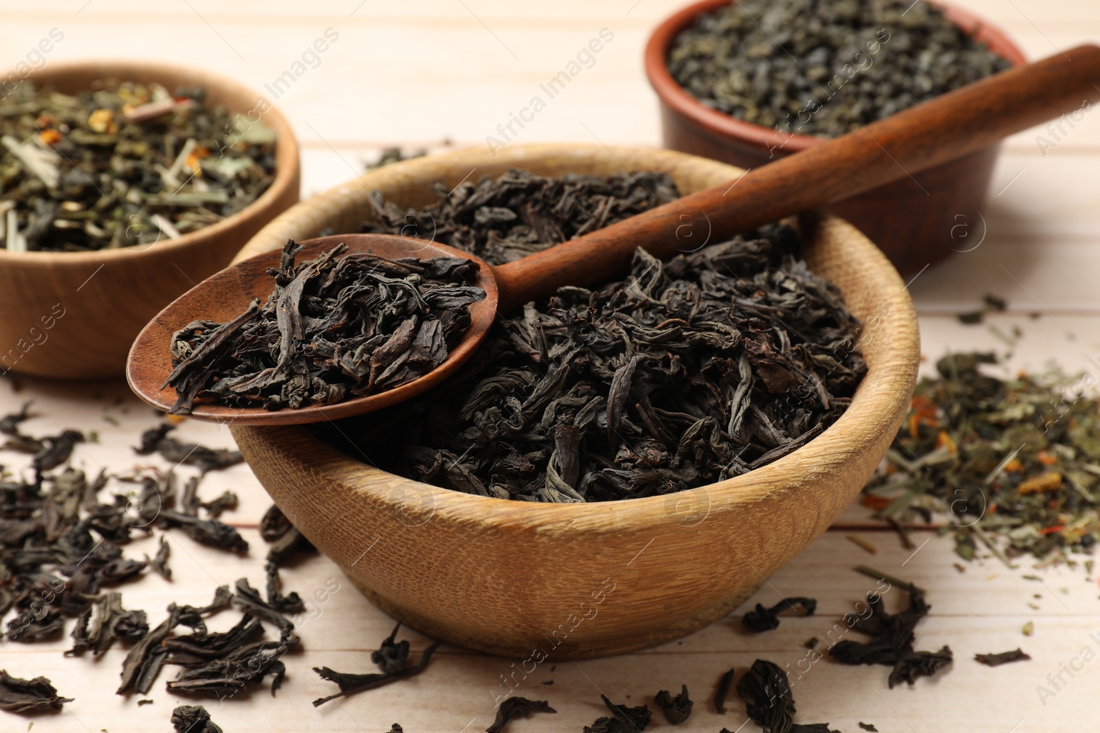 Photo of Different dry tea leaves in bowls and spoon on wooden table, closeup