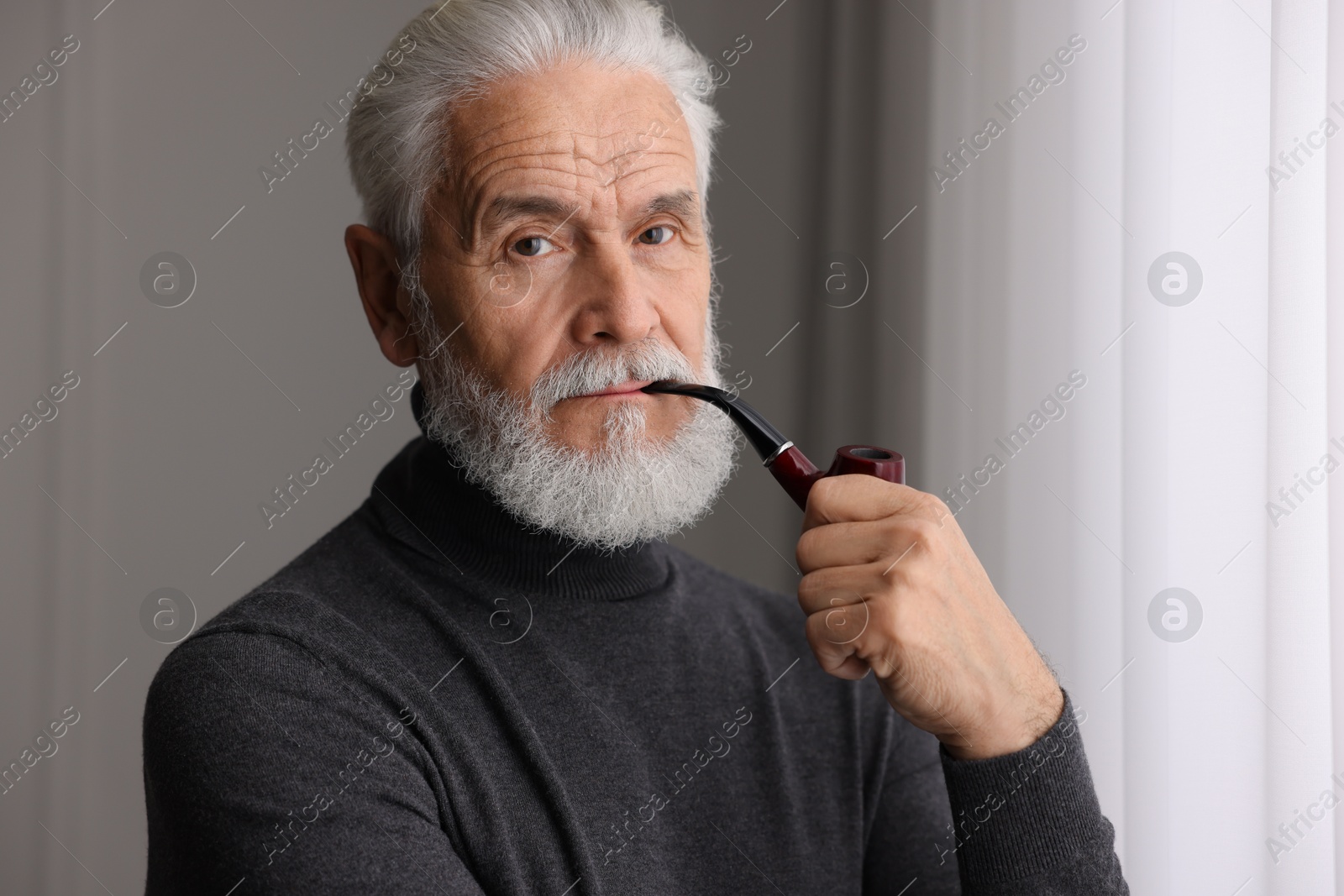 Photo of Portrait of handsome bearded man with tobacco pipe near window indoors