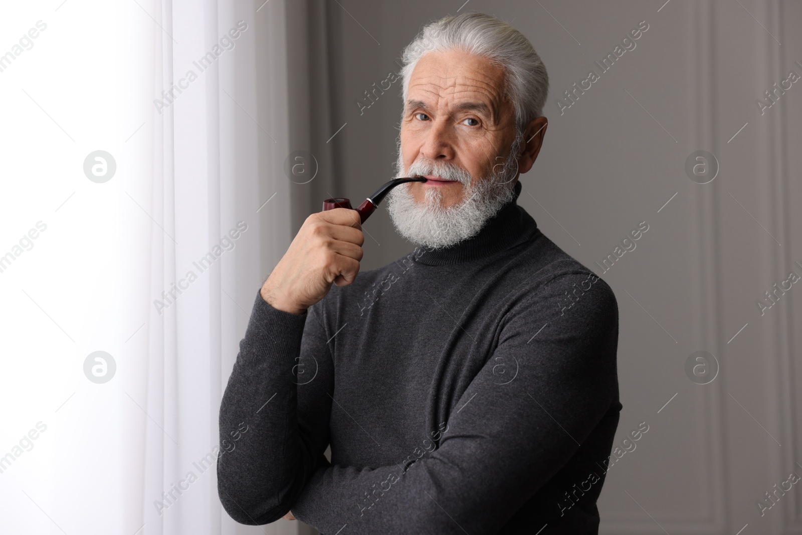 Photo of Portrait of handsome bearded man with tobacco pipe near window indoors