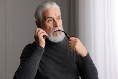 Photo of Portrait of handsome bearded man with tobacco pipe near window indoors