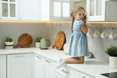 Photo of Little girl sitting on counter and reaching towards shelf in kitchen