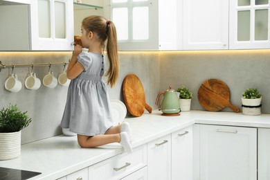 Photo of Little girl sitting on counter and reaching towards shelf in kitchen