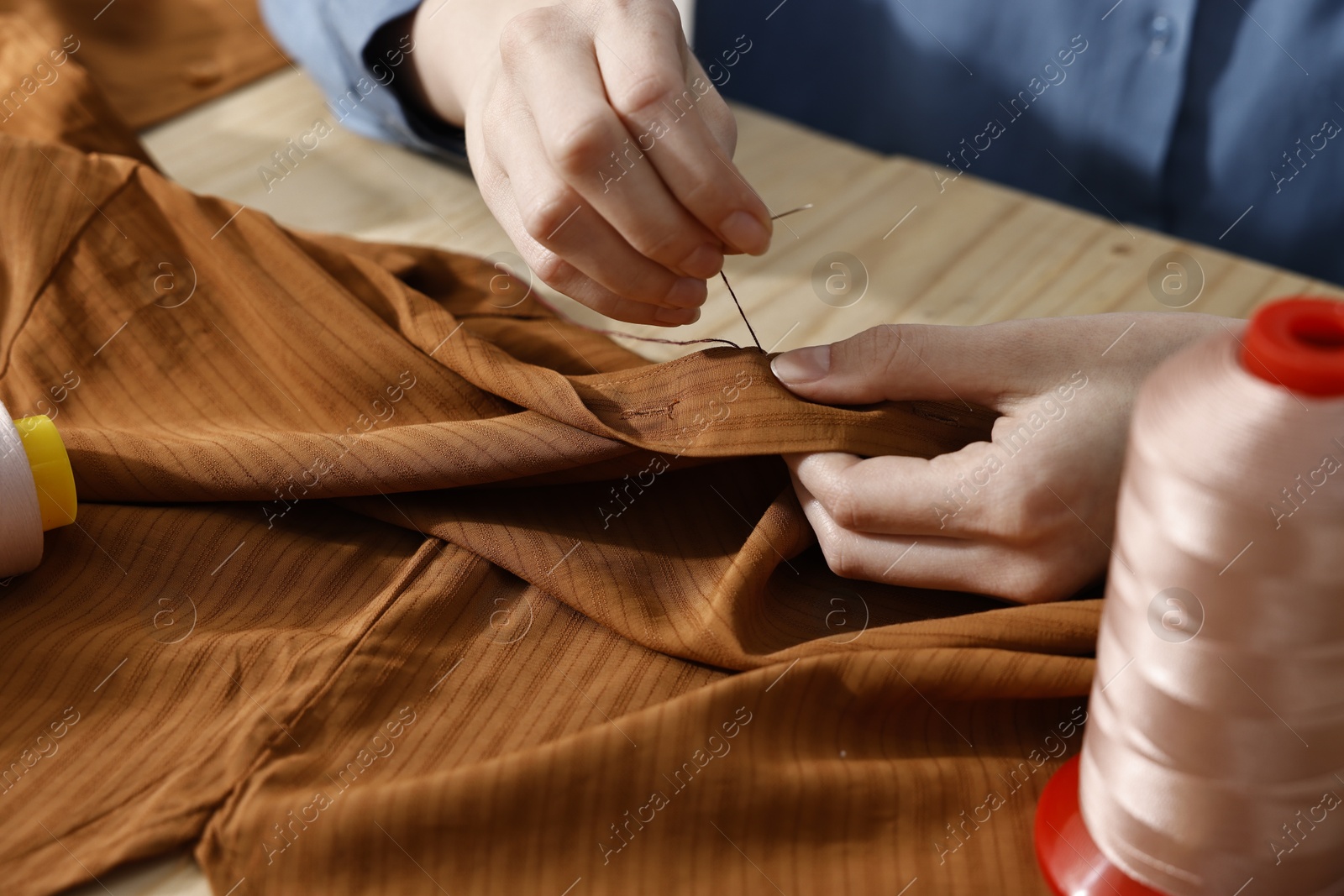 Photo of Woman sewing brown shirt with needle and thread at table, closeup