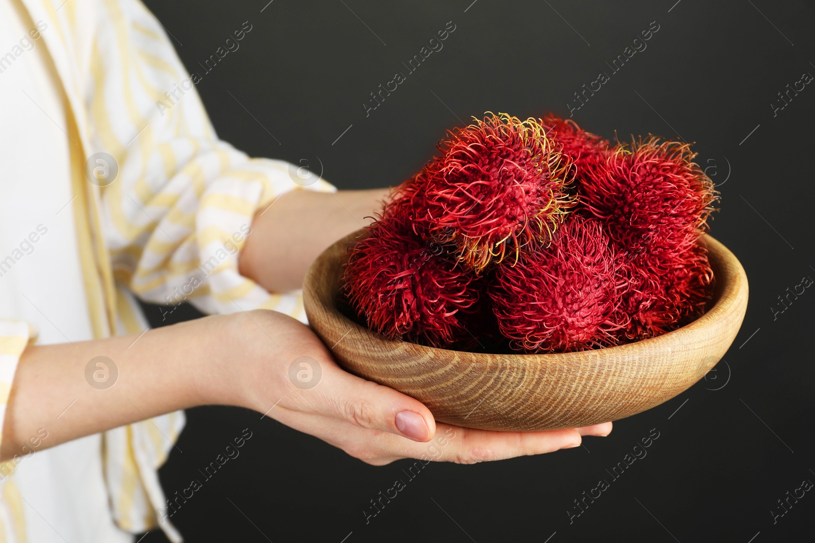 Photo of Woman holding fresh rambutans in bowl on black background, closeup