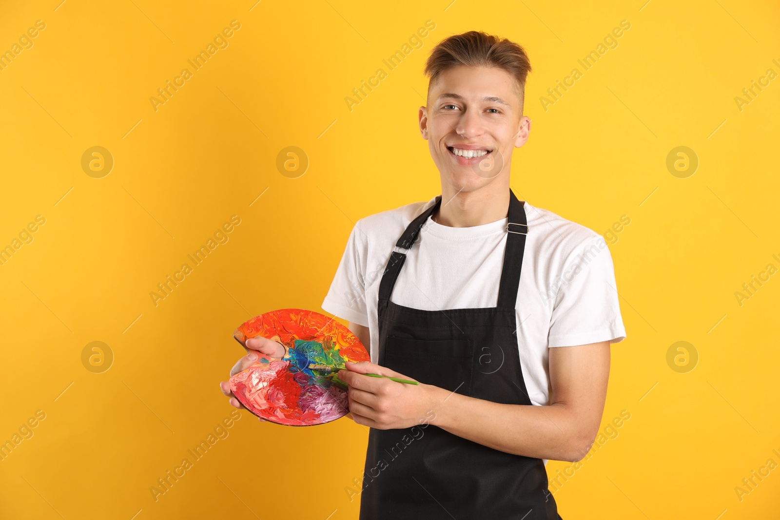 Photo of Smiling man with paintbrush mixing paints on palette against orange background. Space for text