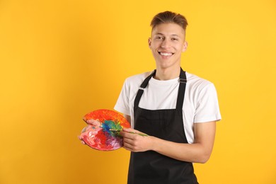 Photo of Smiling man with paintbrush mixing paints on palette against orange background. Space for text