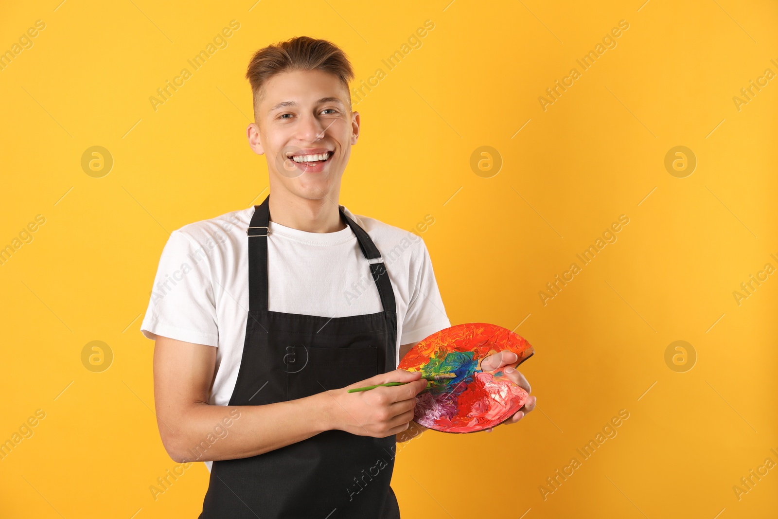 Photo of Smiling man with paintbrush mixing paints on palette against orange background. Space for text