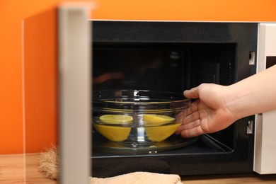 Photo of Woman putting bowl with water and lemon into microwave oven at table indoors, closeup