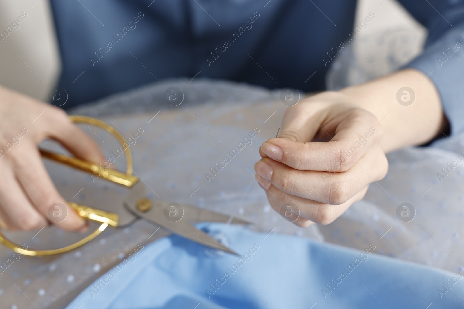 Photo of Woman sewing button with needle and thread onto blue shirt indoors, closeup