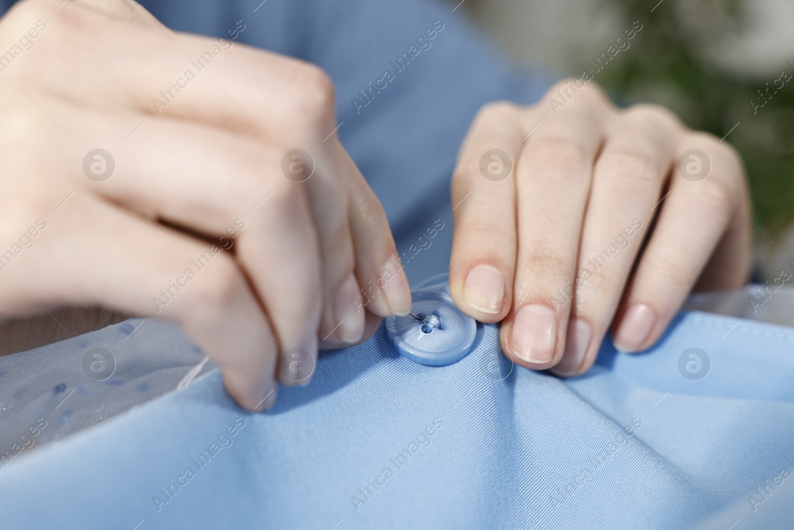 Photo of Woman sewing button with needle and thread onto blue shirt, closeup