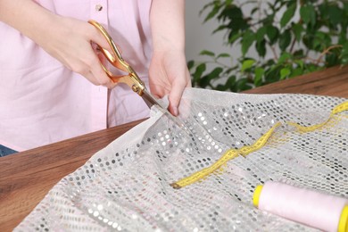 Photo of Woman cutting fabric with sequins at table in workshop, closeup