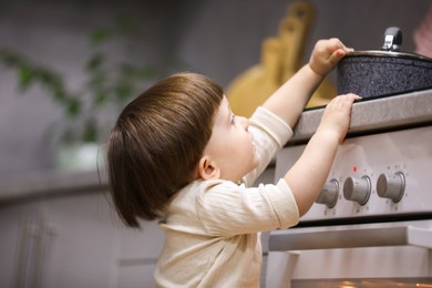 Photo of Little boy playing with pot on stove in kitchen. Dangerous situation