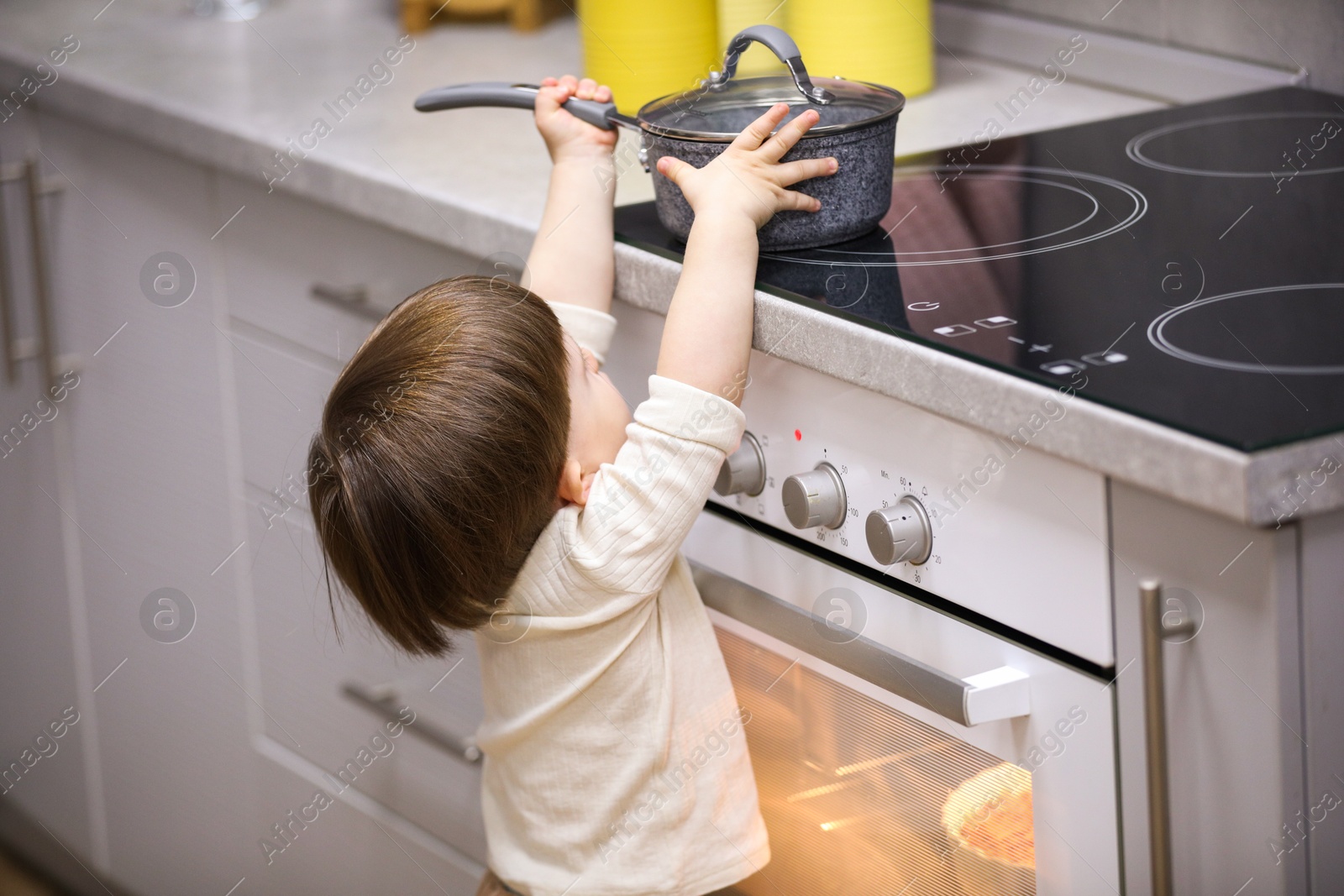 Photo of Little boy playing with pot on stove in kitchen, back view. Dangerous situation