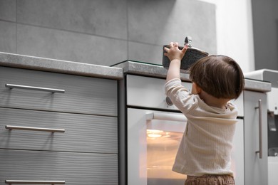 Photo of Little boy playing with pot on stove in kitchen, back view. Dangerous situation