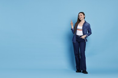 Photo of Smiling woman in stylish jeans pointing upwards on light blue background. Space for text