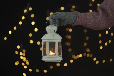 Photo of Woman holding Christmas lantern with burning candle on dark background with blurred lights, closeup. Bokeh effect