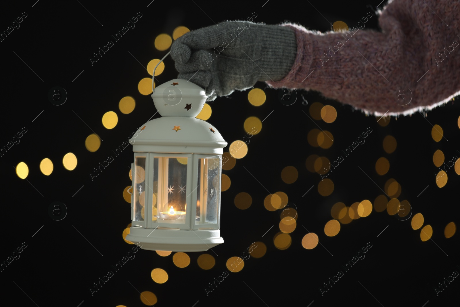 Photo of Woman holding Christmas lantern with burning candle on dark background with blurred lights, closeup. Bokeh effect