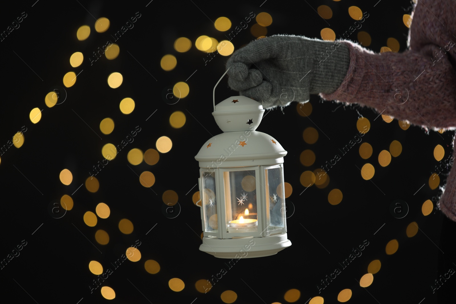 Photo of Woman holding Christmas lantern with burning candle on dark background with blurred lights, closeup. Bokeh effect