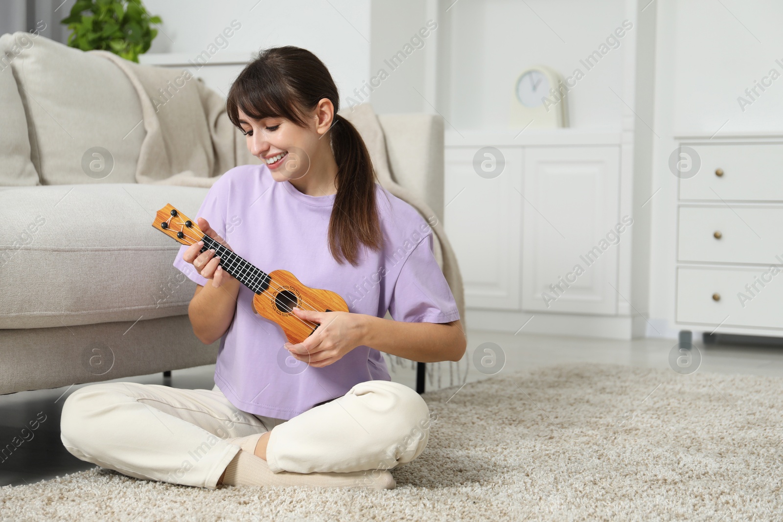 Photo of Happy woman playing ukulele on floor at home, space for text