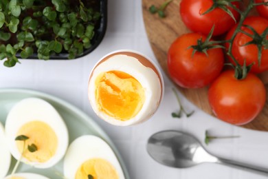 Photo of Boiled eggs, tomatoes and microgreens on white tiled table, flat lay