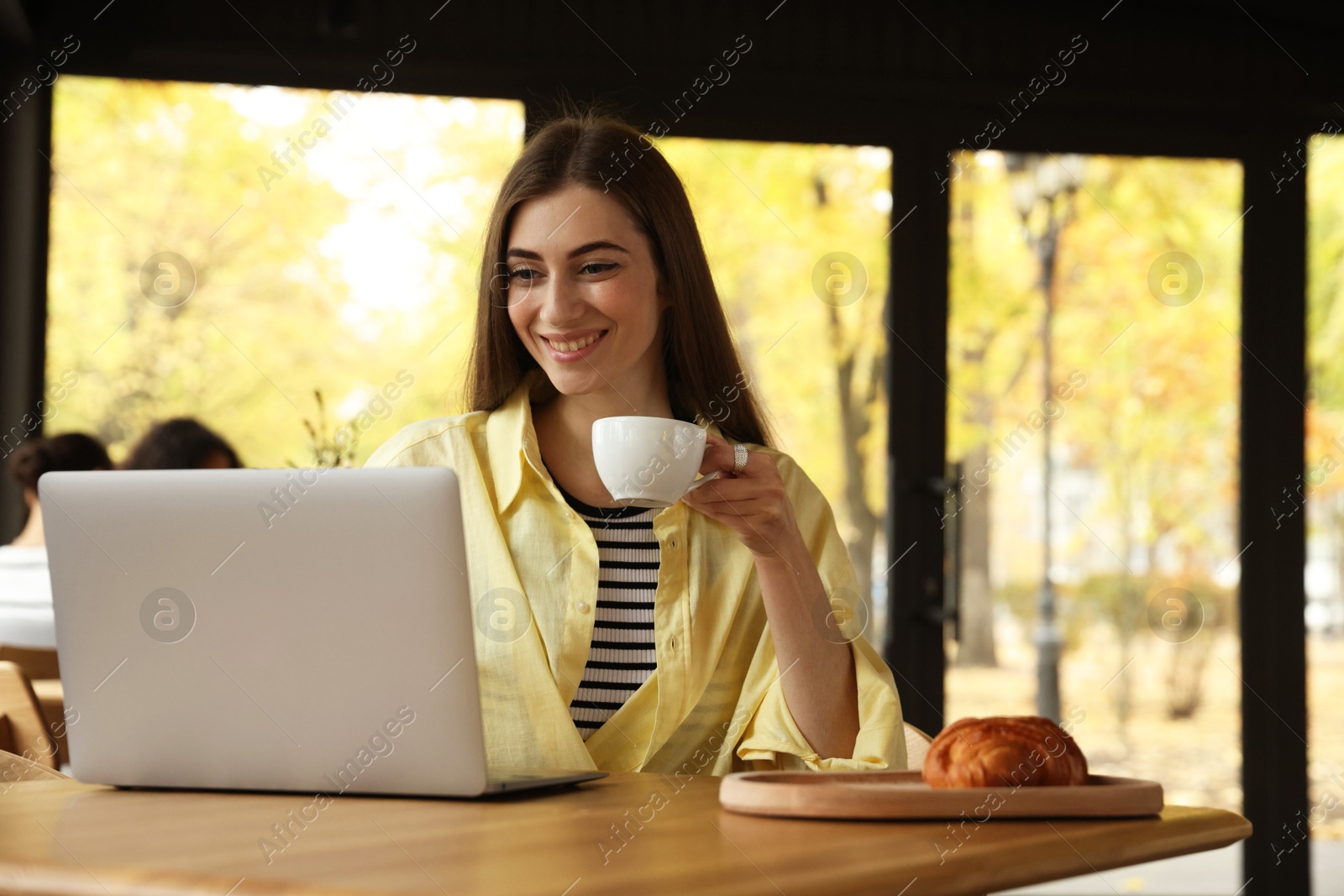 Photo of Woman with coffee and croissant working on laptop at table in cafe