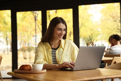 Photo of Woman with coffee and croissant working on laptop at table in cafe