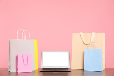 Photo of Internet shopping. Laptop and colorful paper bags on wooden table against pink background