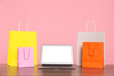 Photo of Internet shopping. Laptop and colorful paper bags on wooden table against pink background