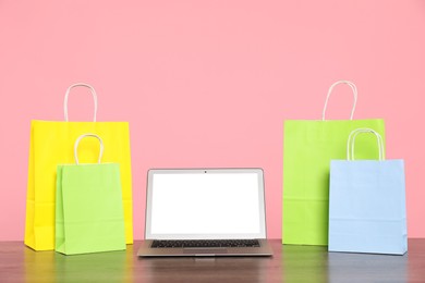 Photo of Internet shopping. Laptop and colorful paper bags on wooden table against pink background