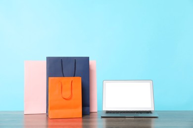 Photo of Internet shopping. Laptop and colorful paper bags on wooden table against light blue background