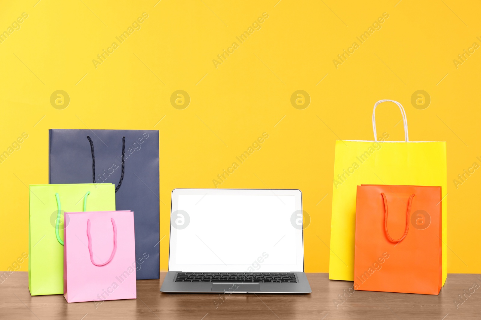 Photo of Internet shopping. Laptop and colorful paper bags on wooden table against yellow background