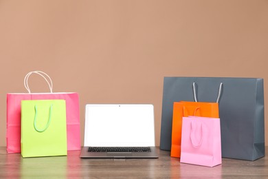 Photo of Internet shopping. Laptop and colorful paper bags on wooden table against brown background