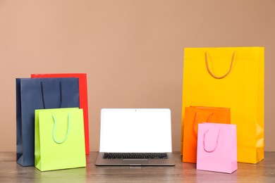 Photo of Internet shopping. Laptop and colorful paper bags on wooden table against brown background