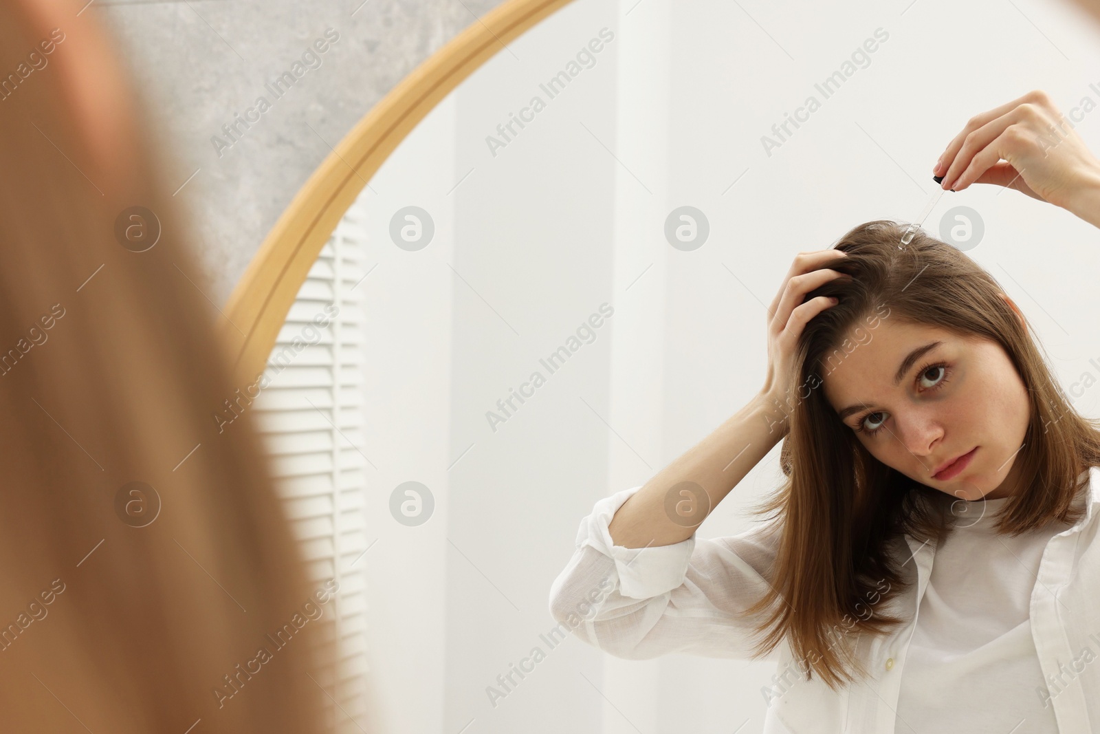 Photo of Hair loss problem. Girl applying serum onto hairline near mirror in bathroom