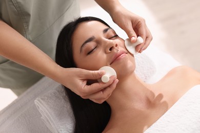 Photo of Young woman receiving facial massage with spa stones in salon