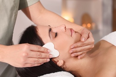 Photo of Young woman receiving facial massage with gua sha tool in spa salon