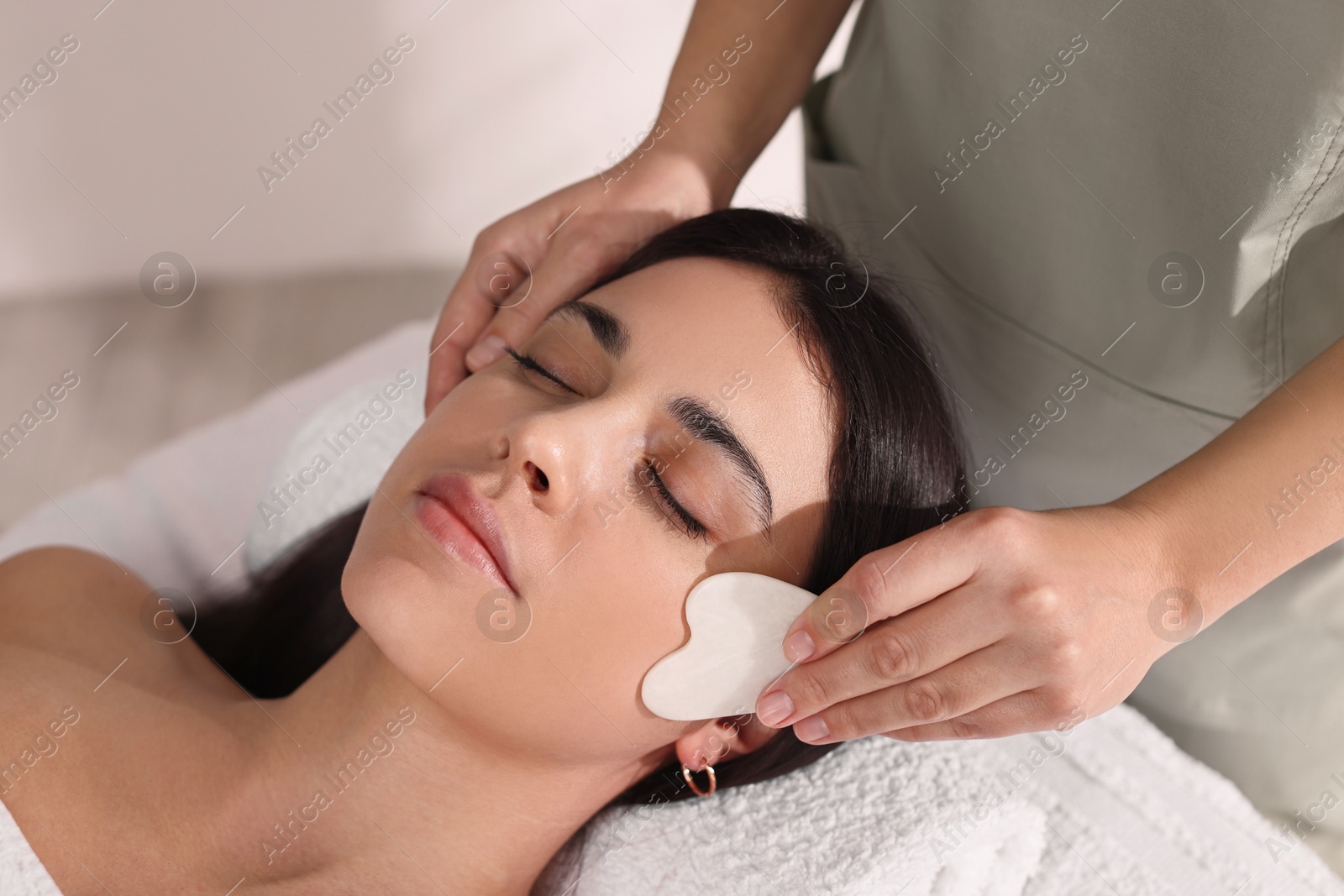 Photo of Young woman receiving facial massage with gua sha tool in spa salon