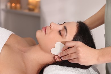 Photo of Young woman receiving facial massage with gua sha tool in spa salon