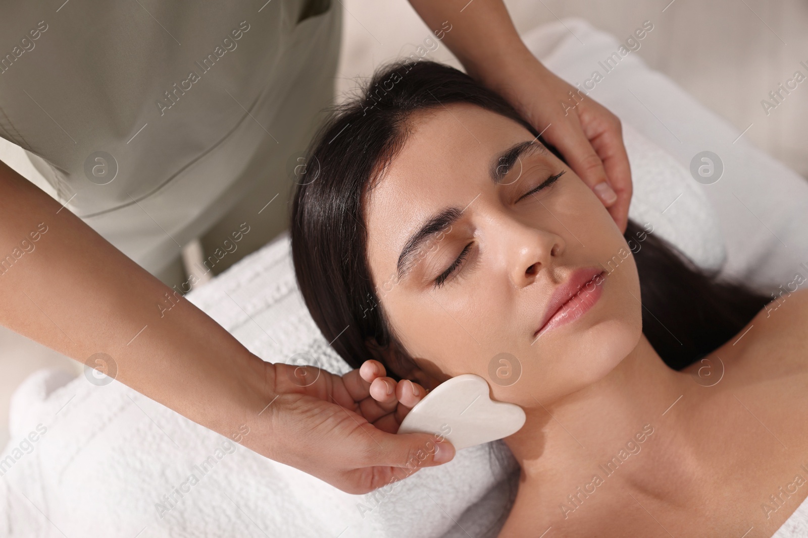 Photo of Young woman receiving facial massage with gua sha tool in spa salon