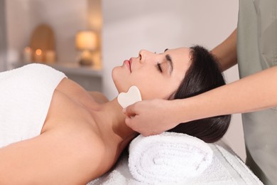 Photo of Young woman receiving facial massage with gua sha tool in spa salon